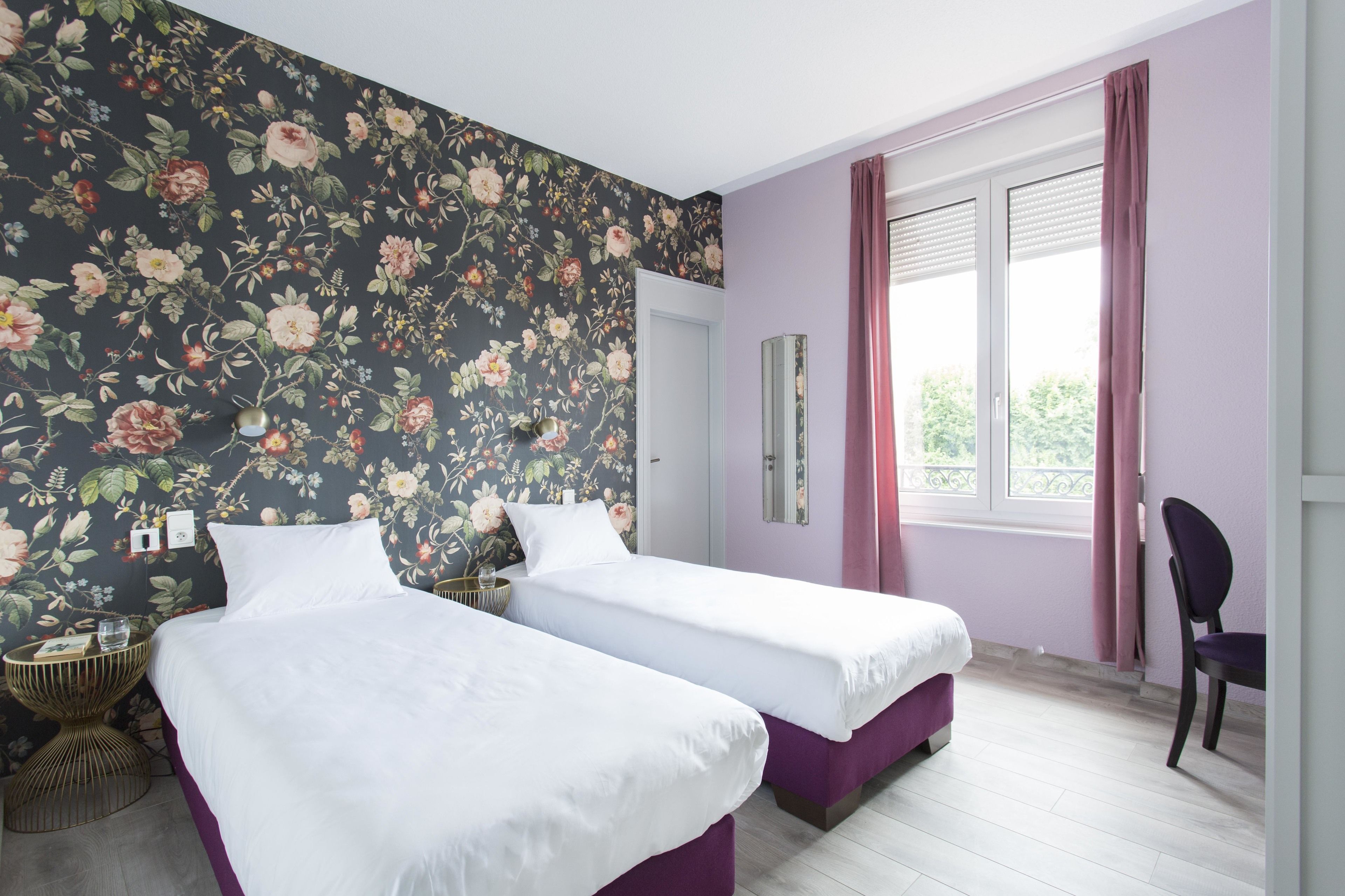 A room with two beds, a chair, a window with curtains, and a floral wallpaper.
