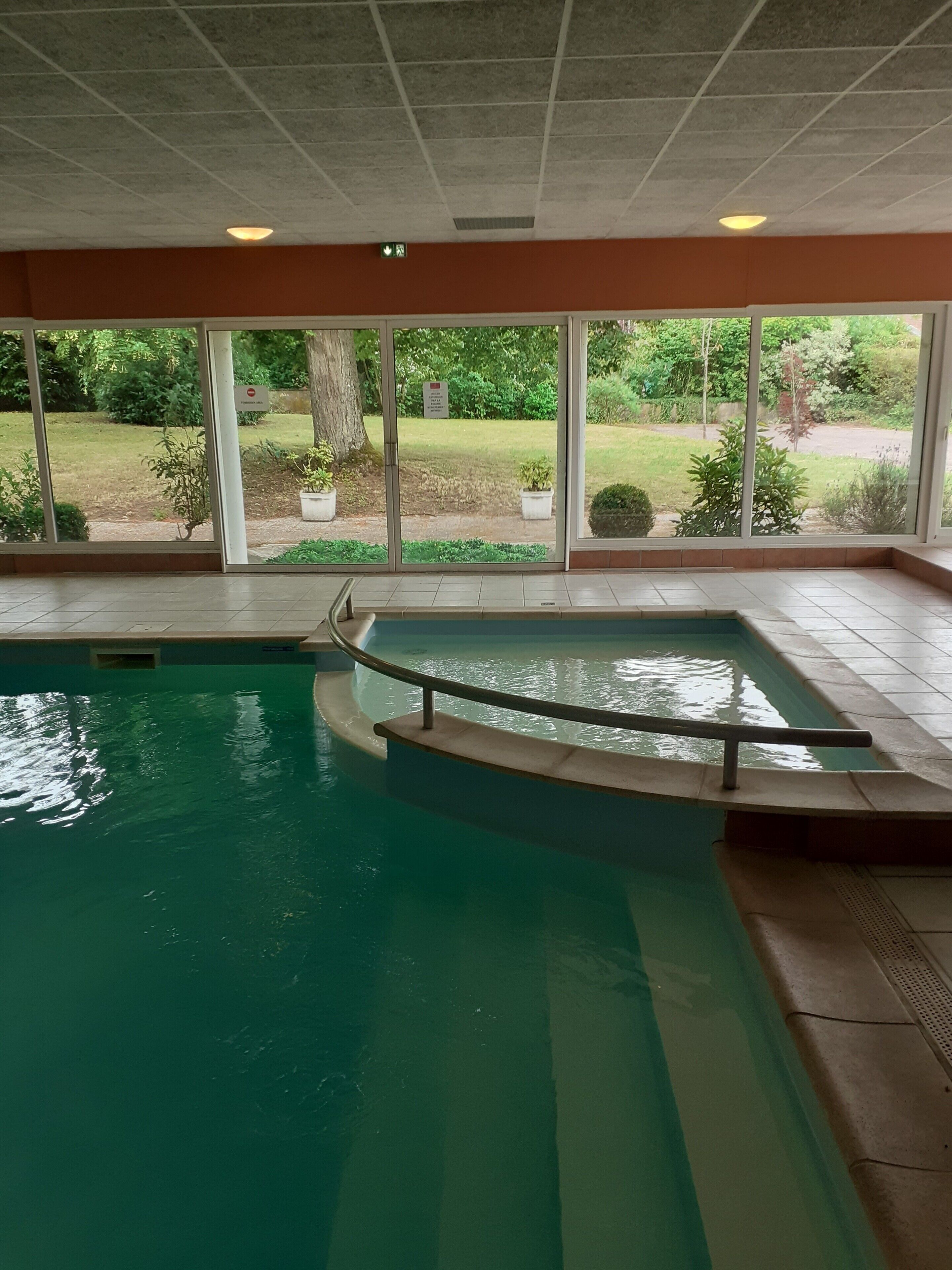 Ein Hallenbad mit großen Glaswänden, einer Metallhandstange und Blick auf einen angelegten Garten.