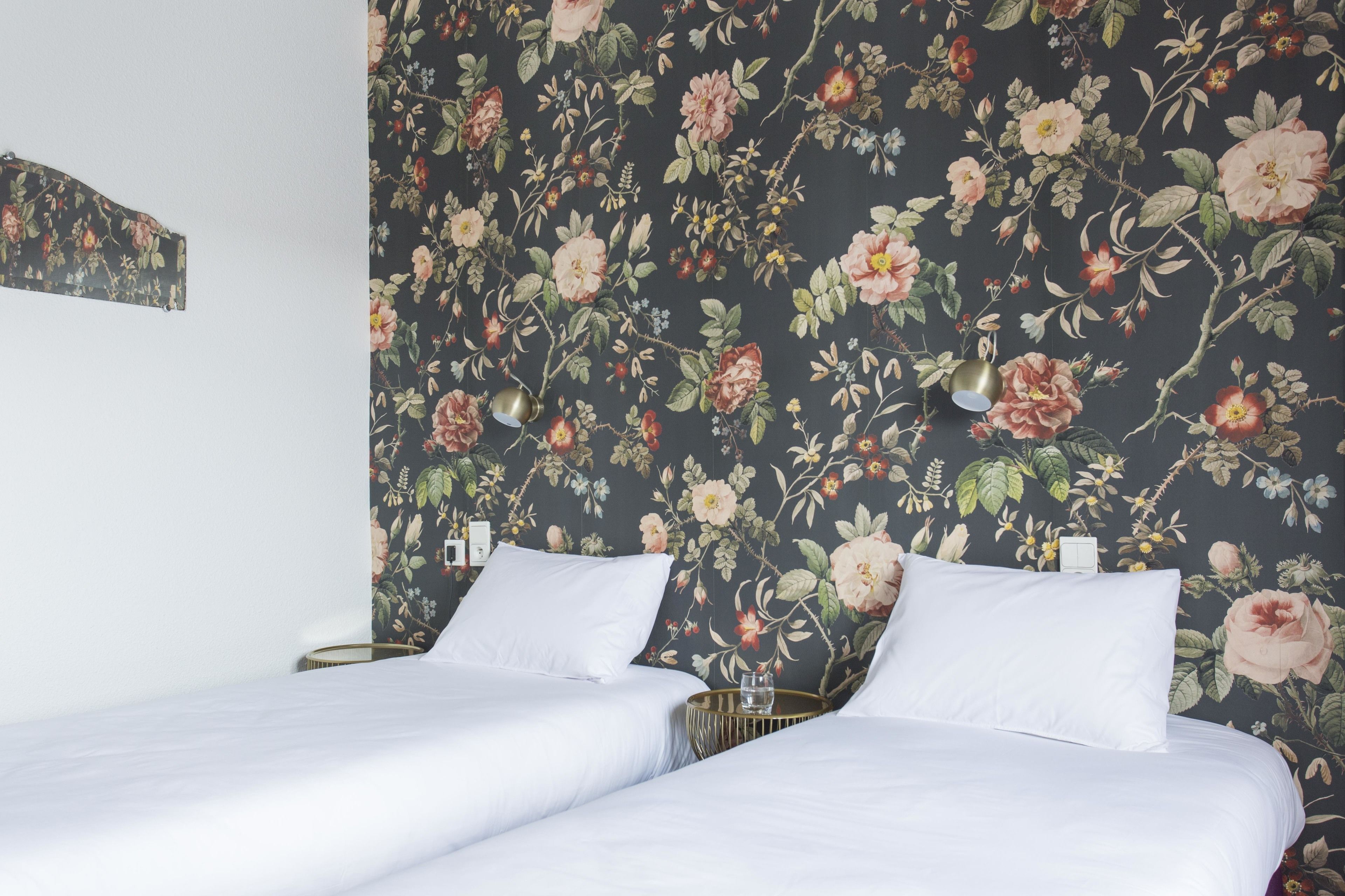 A double bed with white linens, a floral wallpaper, and a bedside table with a lamp.