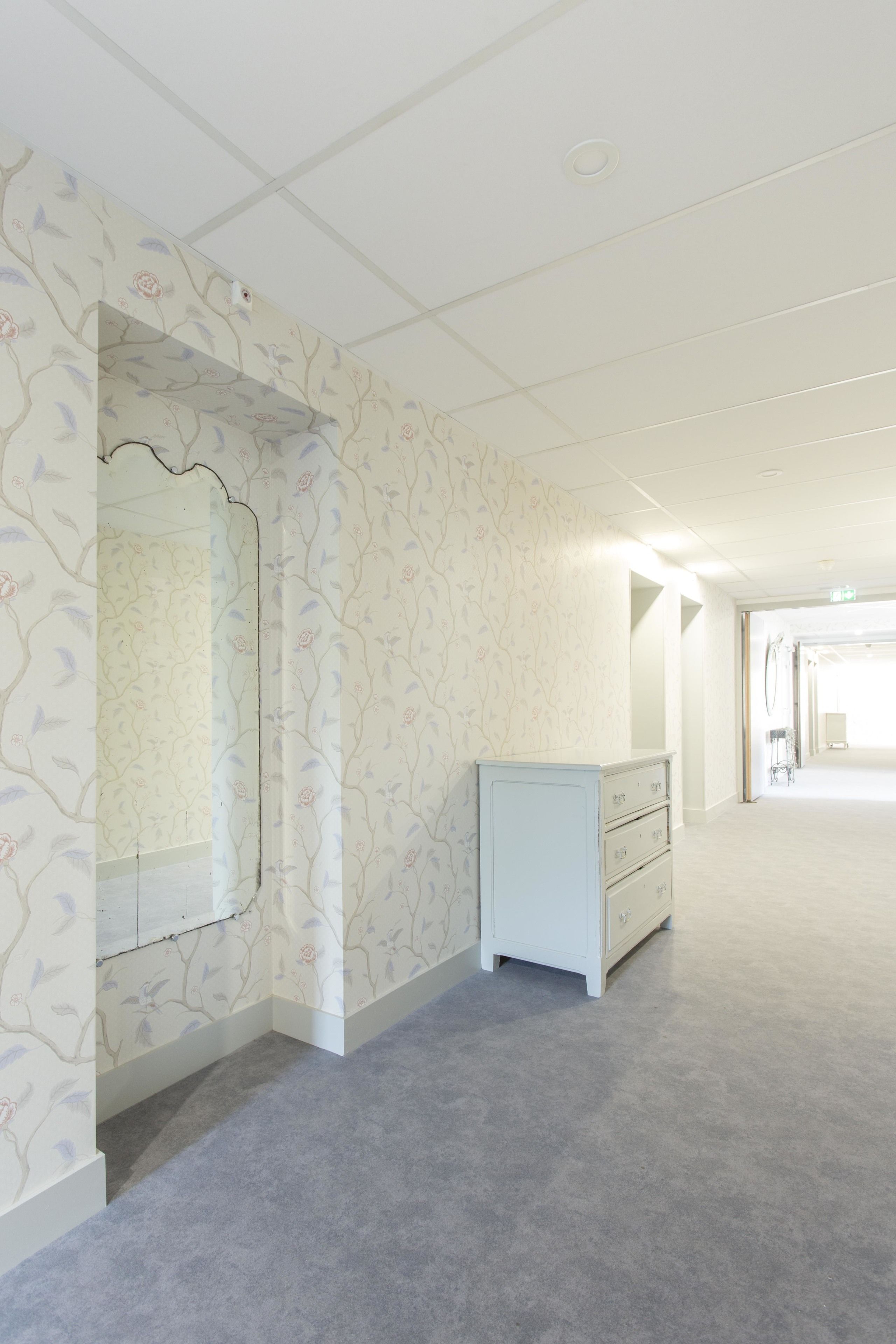 Une chambre d’hôtel avec un papier peint à motifs floraux, une commode blanche et un grand miroir.