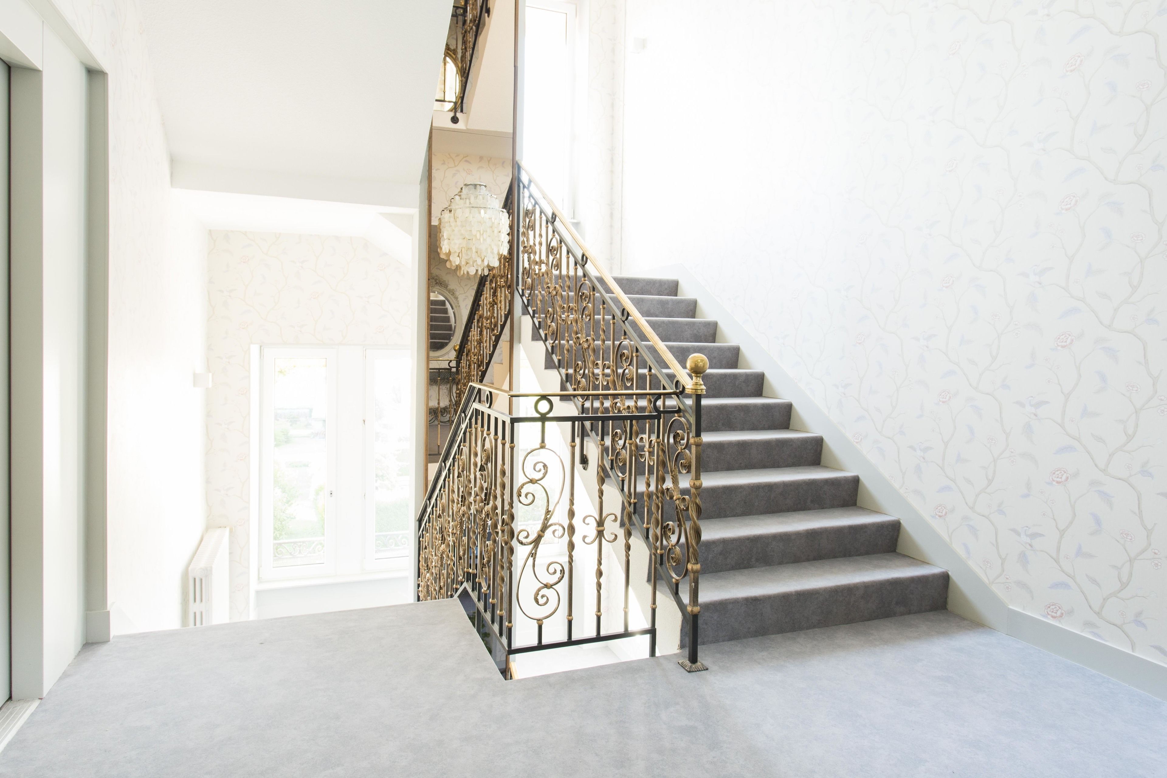 A staircase with ornate black railings and a chandelier, leading to a door with a view of greenery outside.
