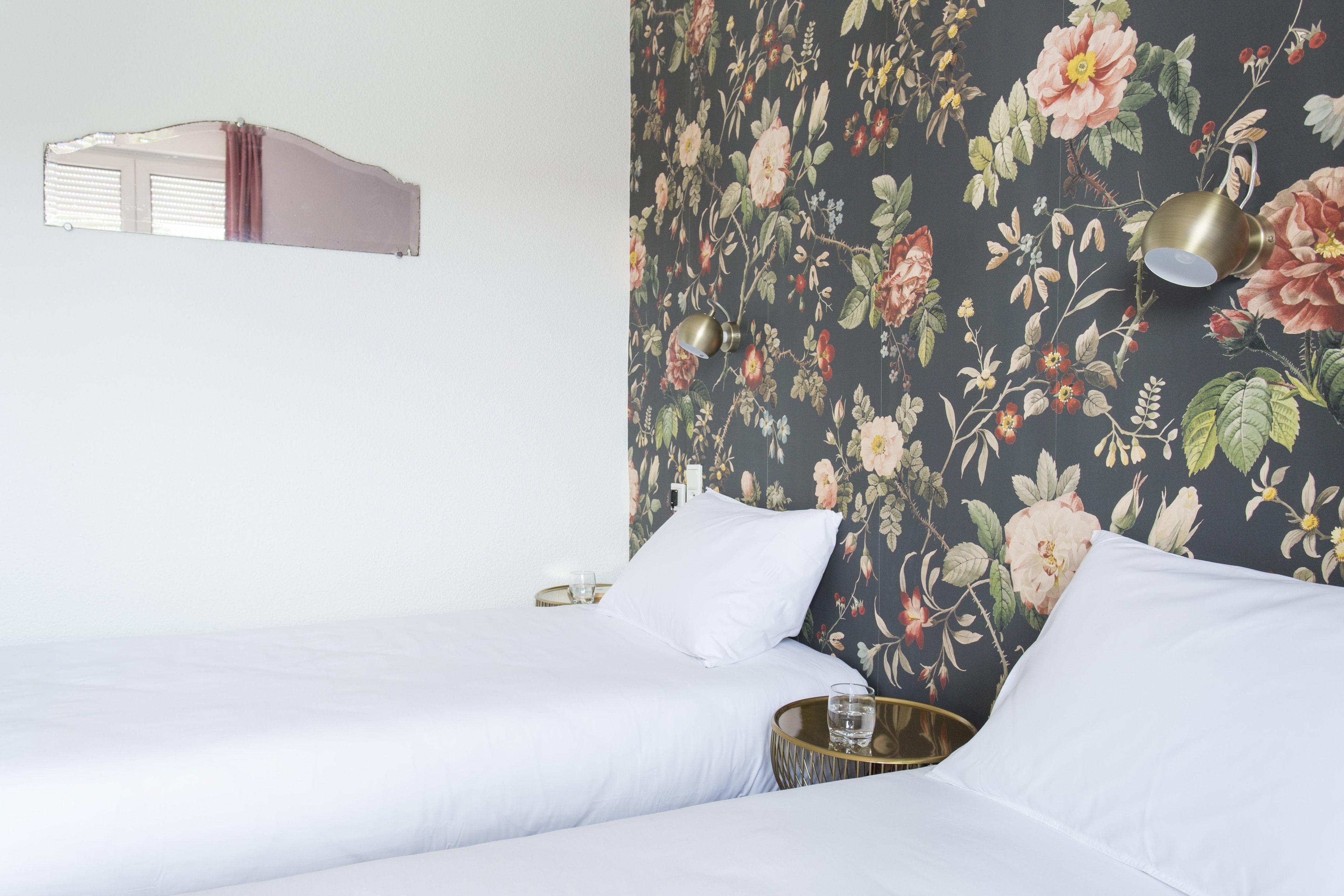 Habitación de hotel con dos camas, papel tapiz floral, una mesita de noche con un vaso y una lámpara de pared.
