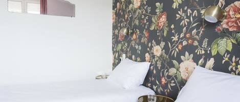 Habitación de hotel con dos camas, papel tapiz floral, una mesita de noche con un vaso y una lámpara de pared.