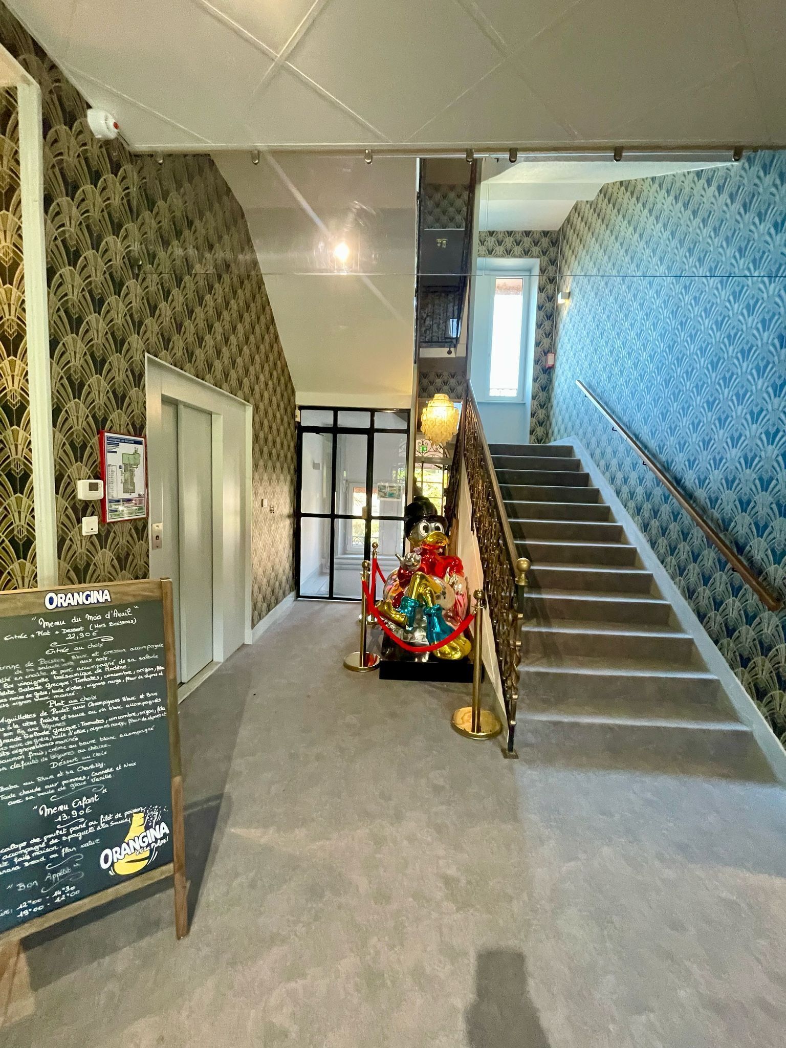 Un couloir d’hôtel avec un escalier, une statue de canard décorative et un panneau affichant le menu.