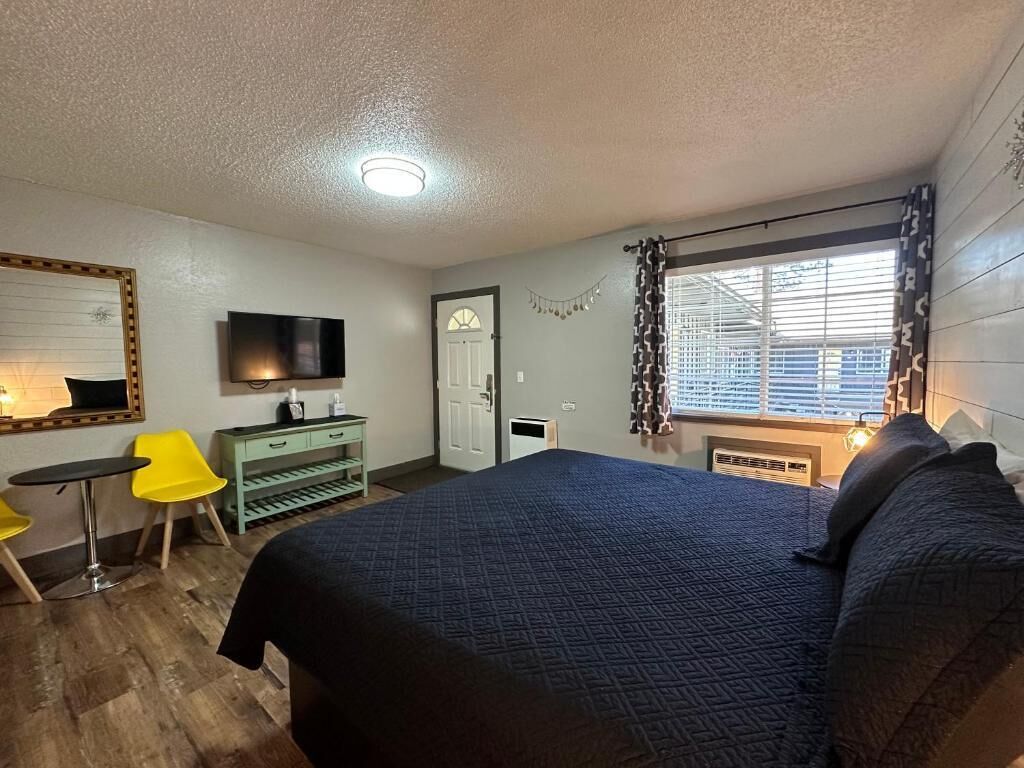 A bedroom with a large bed, a TV mounted on the wall, a desk with a chair, and a window with blinds.