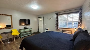 A bedroom with a large bed, a TV mounted on the wall, a desk with a chair, and a window with blinds.