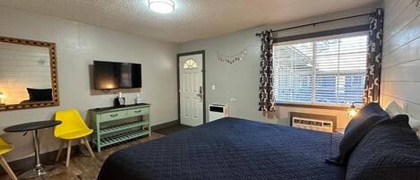 A bedroom with a large bed, a TV mounted on the wall, a desk with a chair, and a window with blinds.
