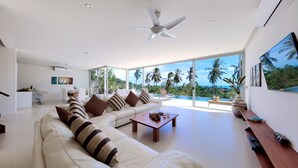 A modern living room with a large sofa, a coffee table, and a TV on a wooden stand. There is a ceiling fan and a view of palm trees and a pool through large windows. - 3 BR Luxury seaview Bang Po-Zoe (Koh Samui)