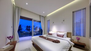 A modern hotel room with a large bed, a bedside table, a nightstand, a window with blinds, and a view of palm trees. - 3 BR Luxury seaview Bang Po-Zoe (Koh Samui)