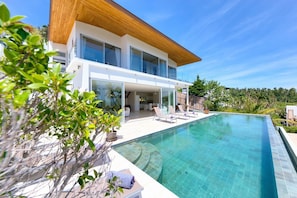 A modern house with a large swimming pool, a wooden deck, and a view of the surrounding greenery.