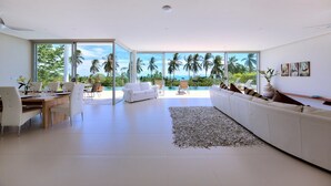 A spacious living room with a large window overlooking a pool and palm trees, a white sofa, and a dining area. - 3 BR Luxury seaview Bang Po-Zoe (Koh Samui)