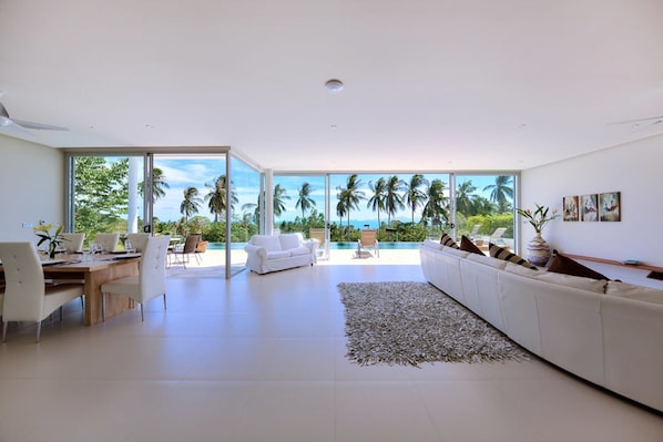 A spacious living room with a large window overlooking a pool and palm trees, a white sofa, and a dining area.