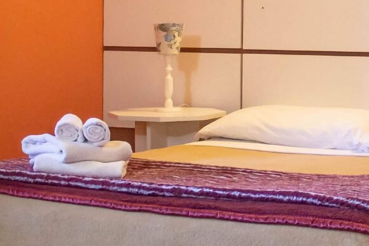 Hotellihuone, jossa on sänky, yöpöytä lampun kanssa ja taulu seinällä.