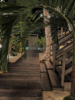 A wooden pathway leading to a gated entrance, surrounded by palm trees and illuminated by string lights.