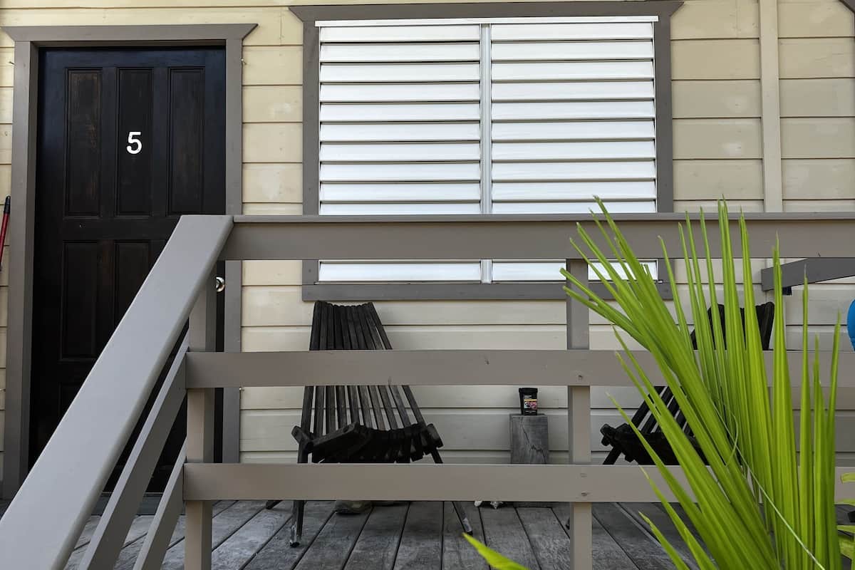 A porch with a black door marked with the number 5, a window with horizontal blinds, a bench, and a small table with a bottle.