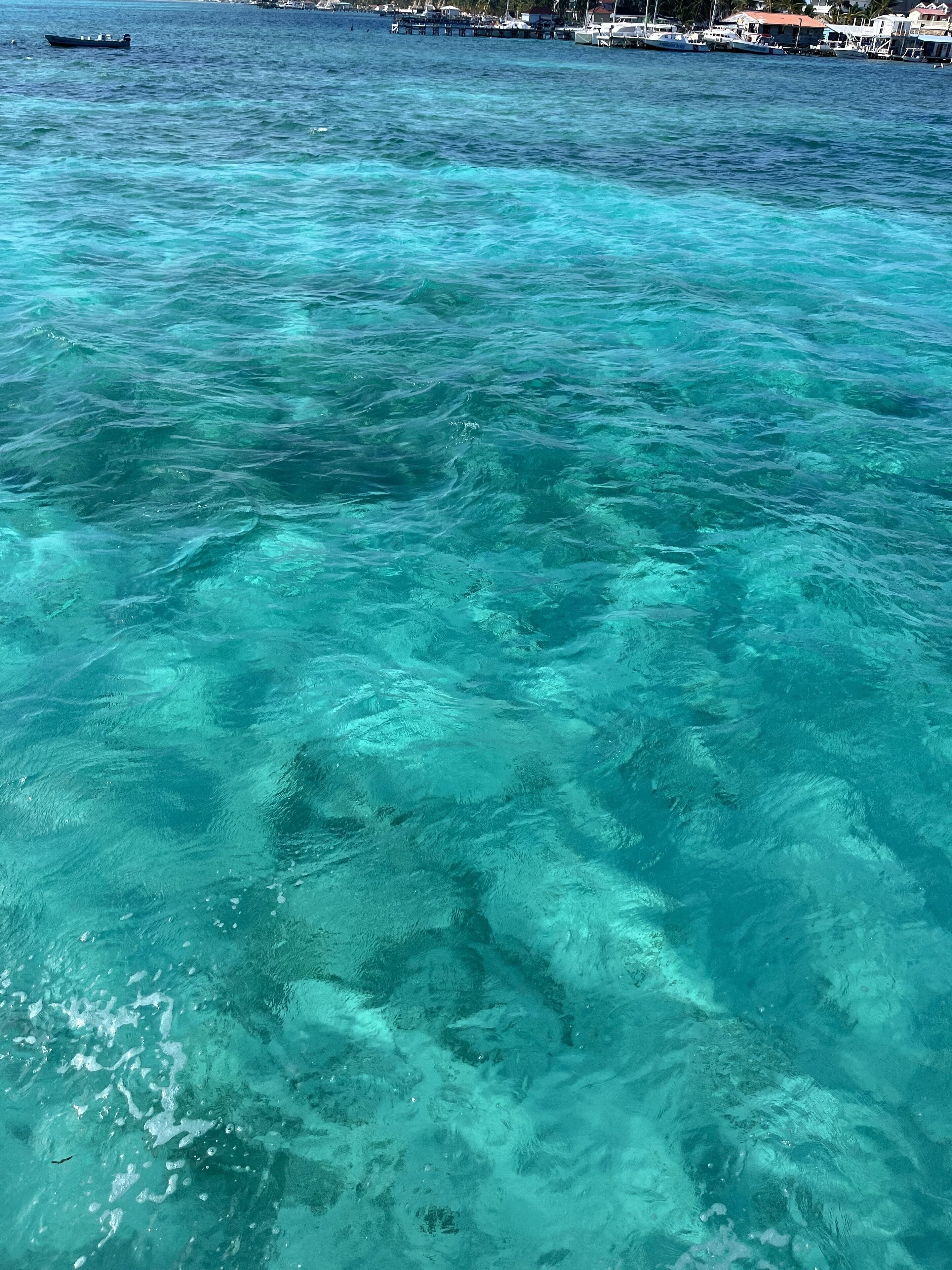 A view of clear turquoise waters with boats and a docked pier in the background.