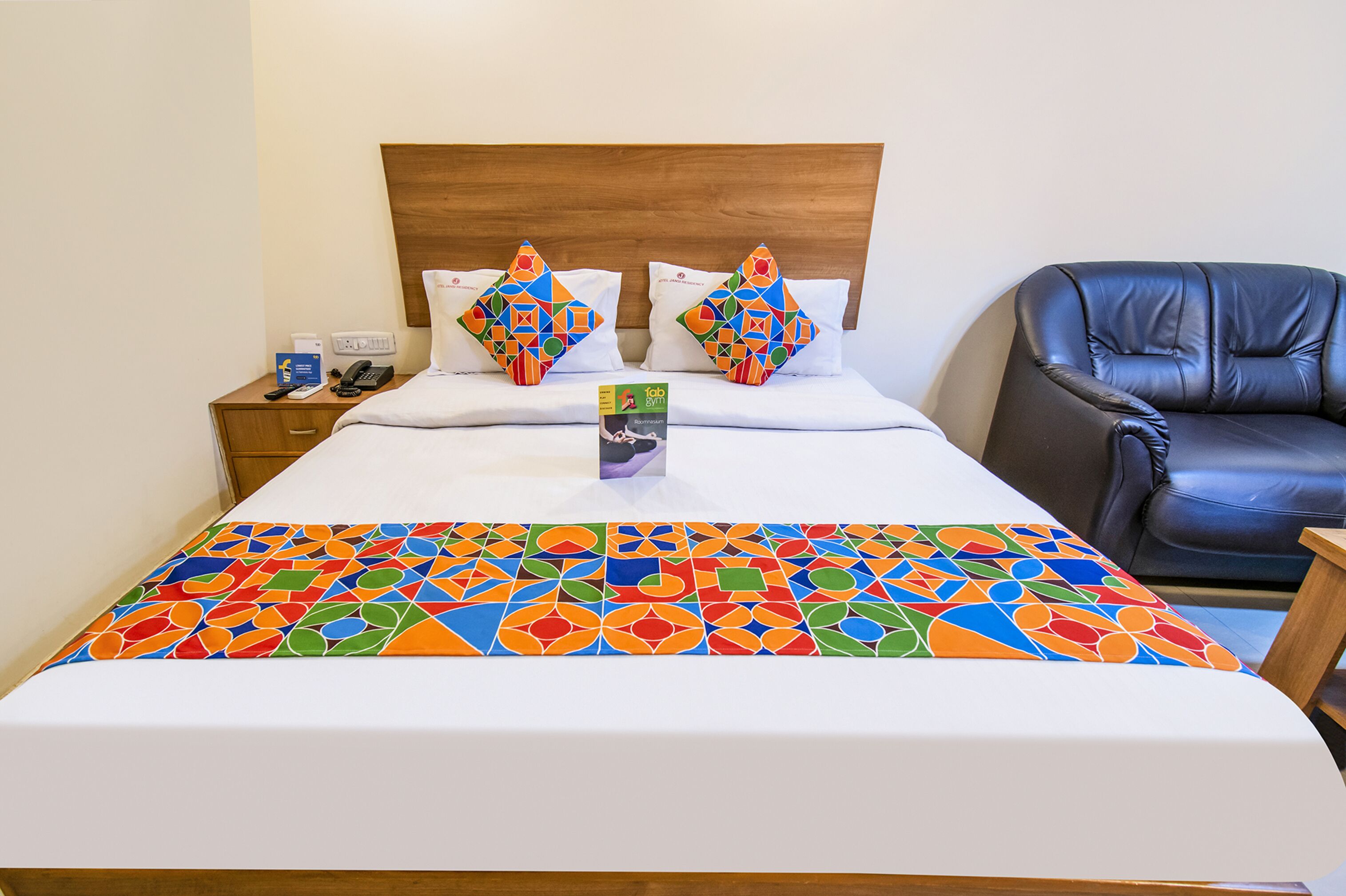 A hotel room with a bed, a wooden headboard, two colorful pillows, a black leather chair, and a bedside table with a phone and a box.