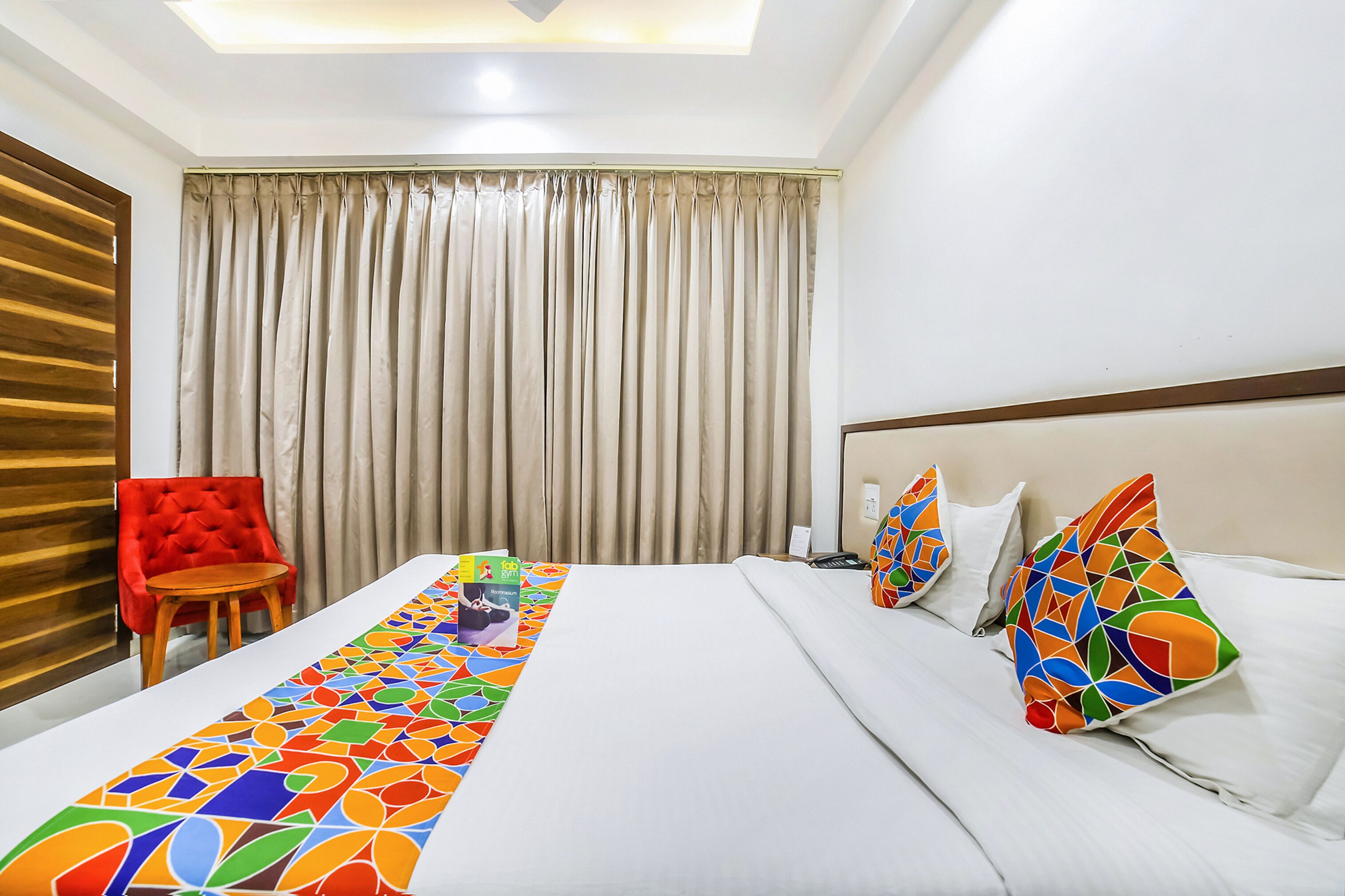 Una habitación de hotel con cama, una silla roja, una cortina y un cojín con un estampado geométrico colorido.