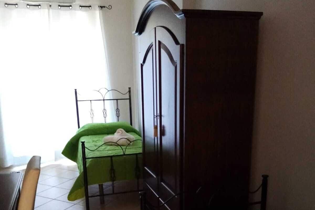 A bedroom with a green bed, a wooden wardrobe, a metal bed frame, and a desk with a chair.