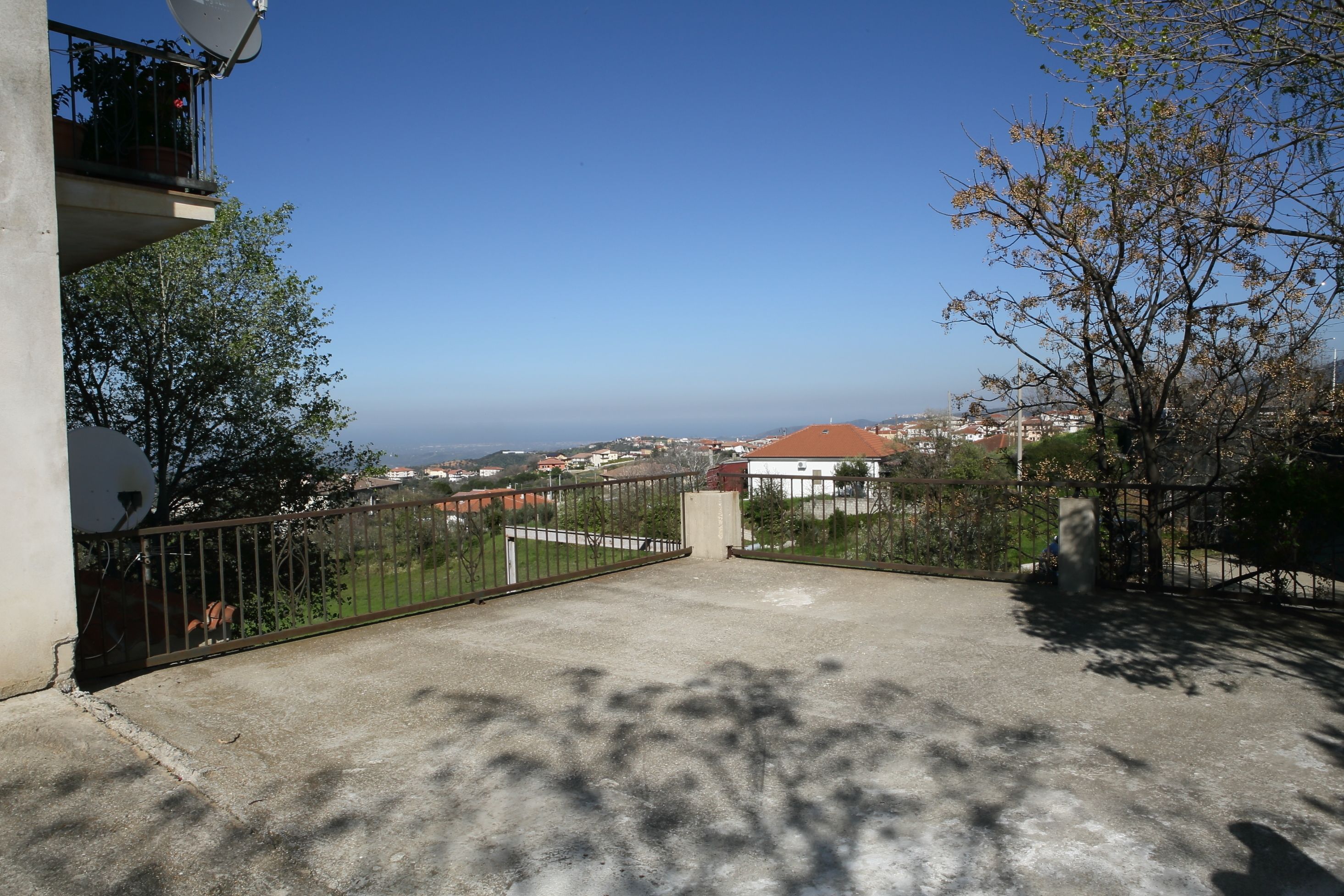 Un balcone in cemento con ringhiera metallica, affacciato su una zona residenziale con case e cielo sereno.