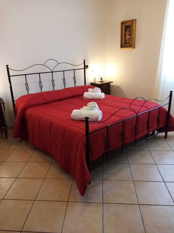 A bed with a red bedspread and three rolled towels on it.