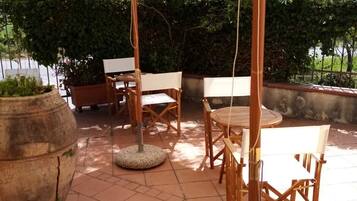 Um pátio externo com mesa e cadeiras redondas, um vaso grande e uma pérgula com cobertura branca.