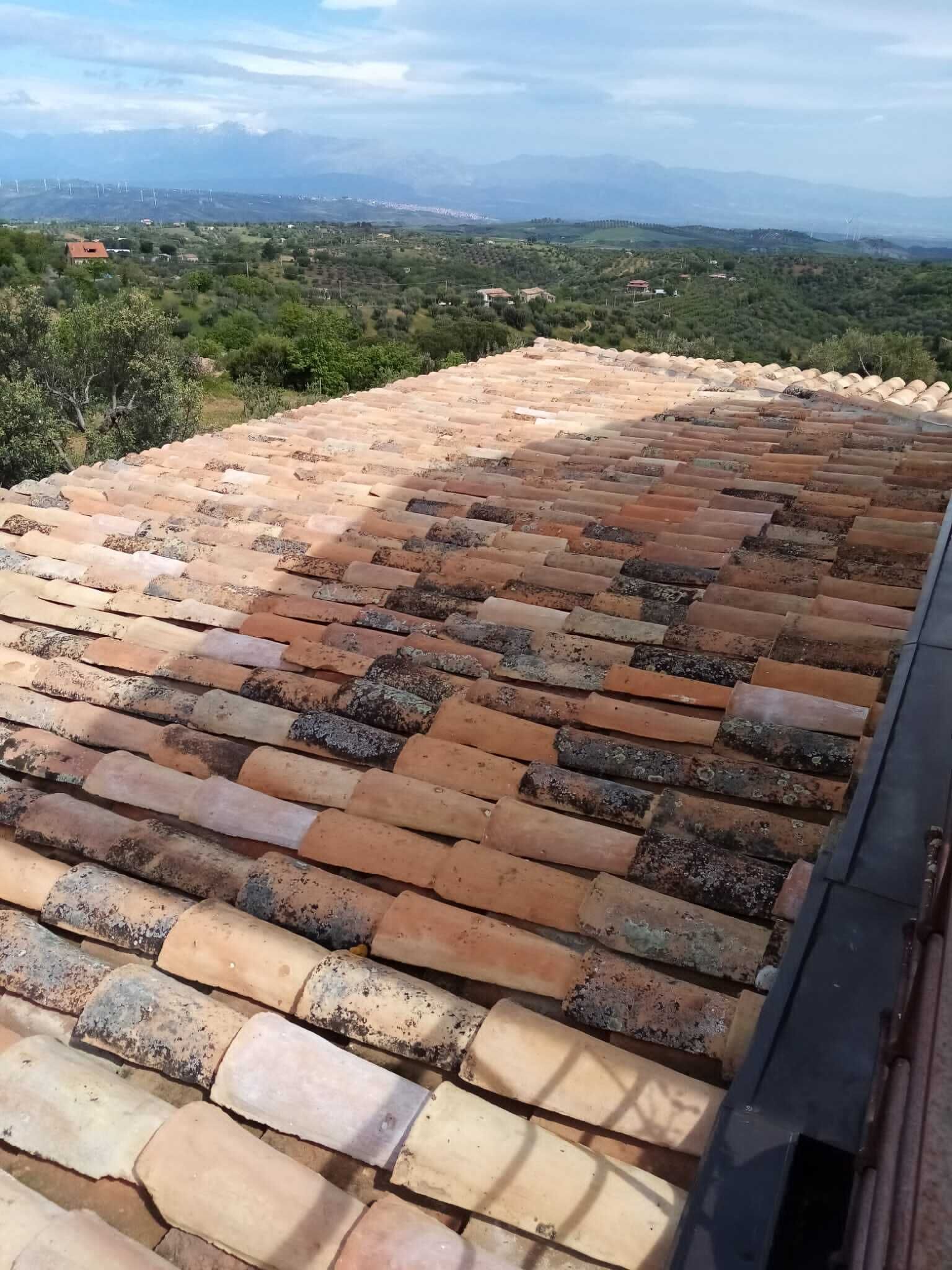 A rooftop with terracotta tiles, overlooking a green landscape with distant buildings and wind turbines.