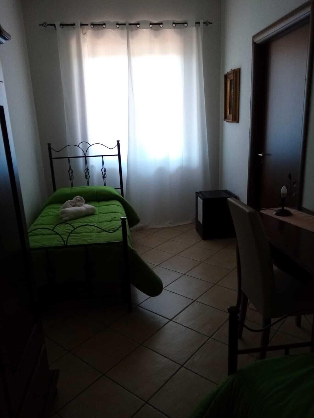 Ein kleiner, schwach beleuchteter Raum mit einem Einzelbett, einem Schreibtisch und einem Stuhl.