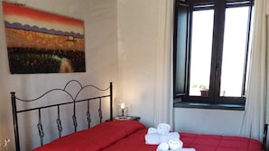 A neatly made bed with a red bedspread and white towels, a nightstand with a lamp, a painting on the wall, and a window with curtains. - La Giara B&B (San Demetrio Corone)