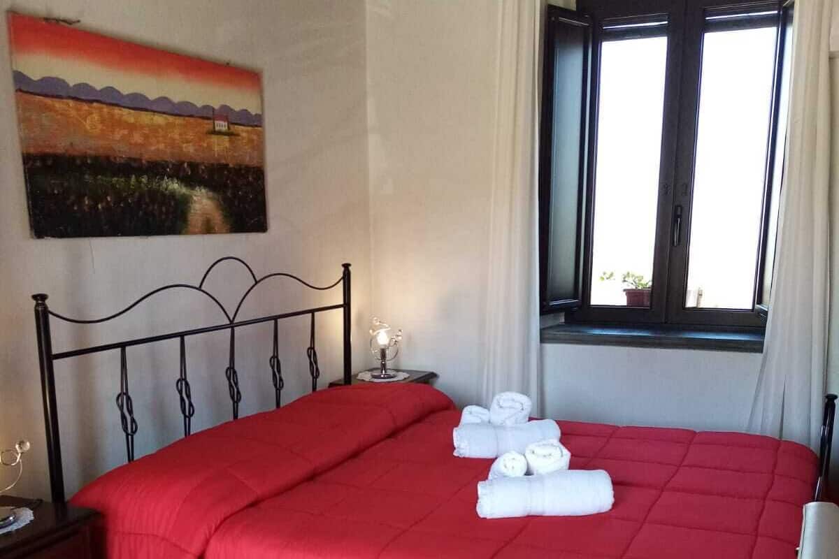 A neatly made bed with a red bedspread and white towels, a nightstand with a lamp, a painting on the wall, and a window with curtains.