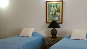 Dos camas individuales con ropa de cama azul y almohadas blancas, una mesita con lámpara y un cuadro de flores en la pared.