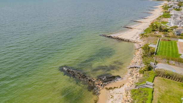 Eine Luftaufnahme einer Küstenregion mit einem breiten, hellen Strand, einer felsigen Küstenlinie und Wohnhäusern direkt am Ufer.