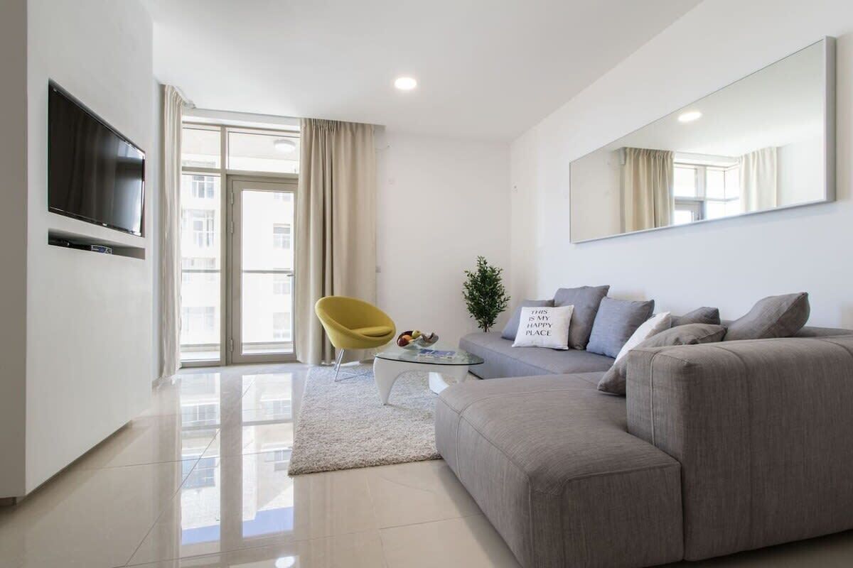 Un salon moderne avec un canapé gris, un fauteuil jaune, une petite table basse et un téléviseur à écran plat fixé au mur.