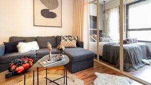 A modern living room with a grey sofa, a coffee table with a globe, and a fruit bowl. There is a large window with curtains, a chandelier, and a bed with a grey duvet.