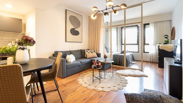 A modern living room with a sofa, dining table, and chairs. There is a large window with curtains, a floor lamp, and a painting on the wall.