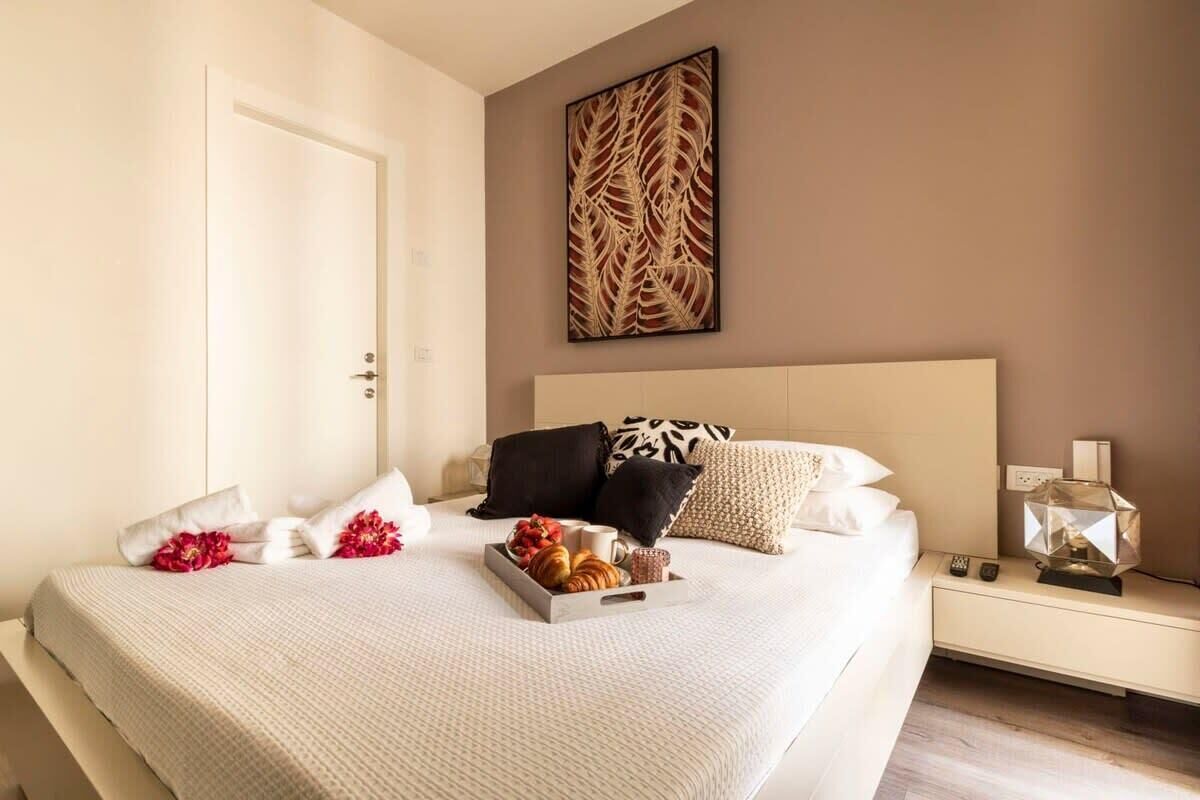 Una cama bien hecha con una bandeja de pasteles y flores, una mesita de noche con una lámpara y una obra de arte en la pared.