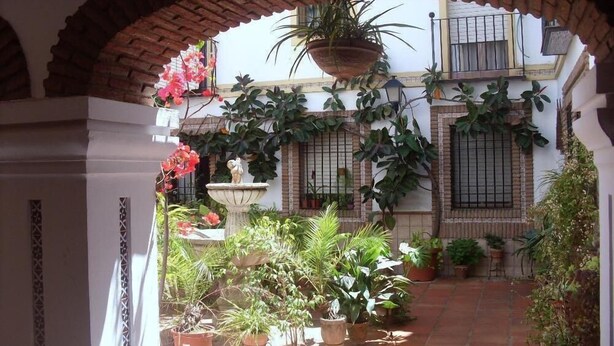 Un patio exterior con un arco de ladrillo, plantas en macetas, una fuente y un edificio con ventanas con rejas.