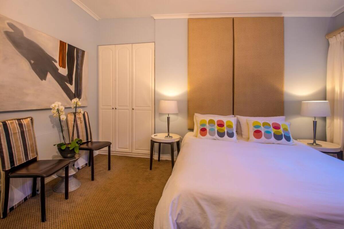 A hotel room with a large bed, a desk, a chair, a wardrobe, and a painting on the wall.