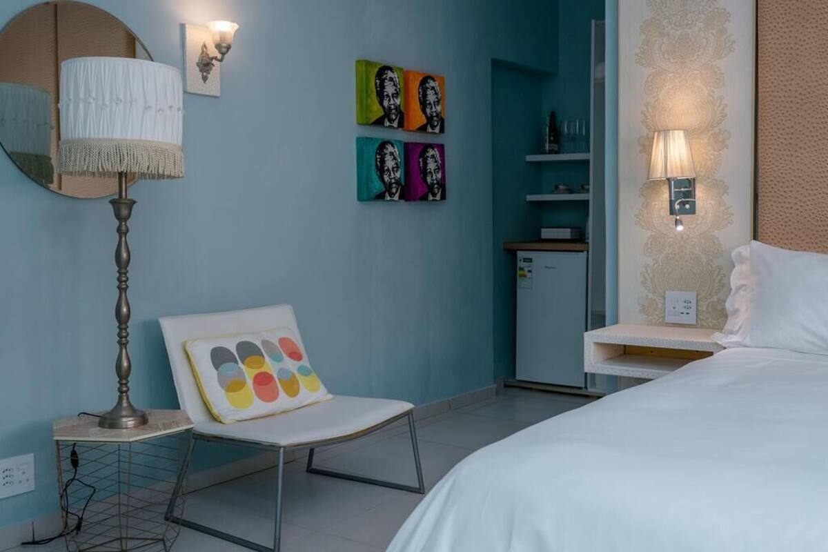 A hotel room with a bed, a chair with a colorful cushion, a lamp, a mirror, and wall art.