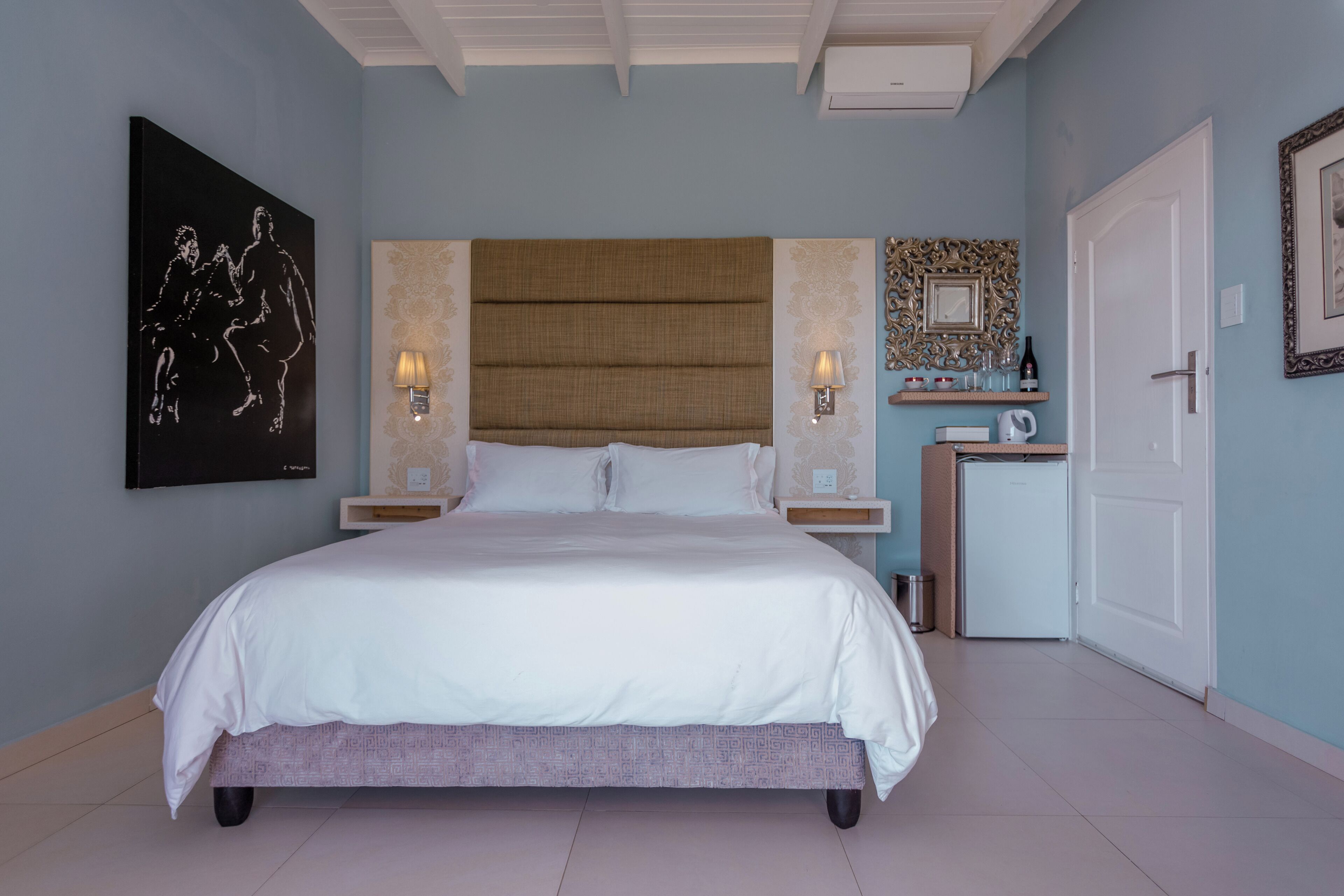 A bedroom with a large bed, two bedside lamps, a nightstand, a wall-mounted TV, a decorative mirror, and a door.