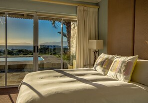 A hotel room with a large bed, a view of a pool and mountains, and a sliding glass door. - Benguela (De Kelders)