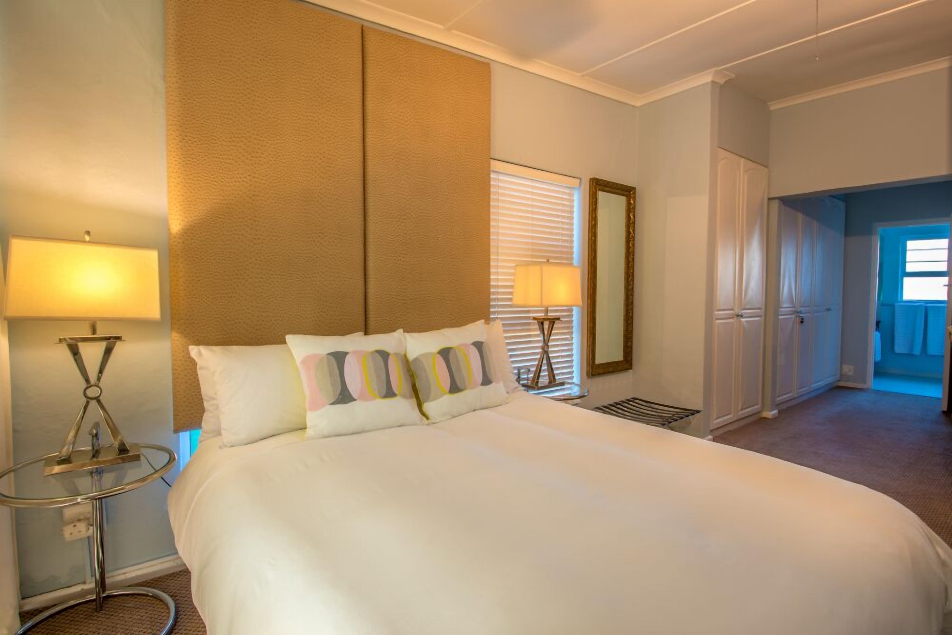 A hotel room with a large bed, a bedside table with a lamp, a mirror, and a window with blinds.