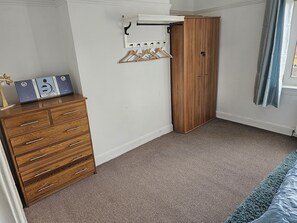 Quarto com cômoda de madeira, guarda-roupa, cabideiro com ganchos e cama com cobre azul.