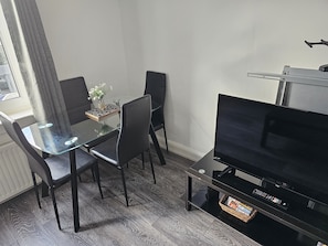 Um espaço de estar compacto com mesa de jantar de vidro, cadeiras pretas, televisão de tela plana e uma pequena mesa lateral.