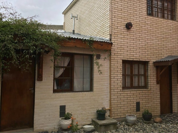 A two-story building with a brick facade, a wooden door, and a window with curtains. There are potted plants on the ground.
