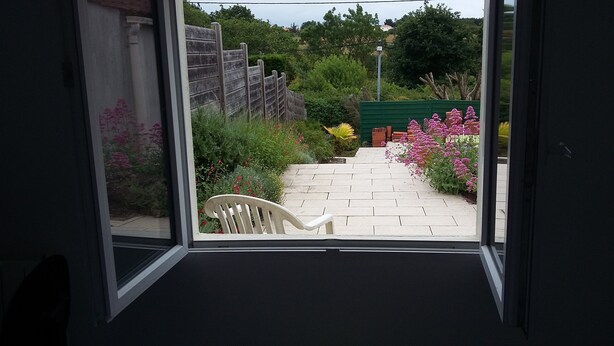 A view through a window of a garden with a paved path, a wooden fence, and various plants and flowers. - 2 minutes walk from the beach (Pornic)