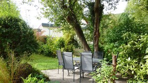 Une terrasse avec une table et des chaises, entourée d’une végétation luxuriante et bordée d’un chemin de pierre.