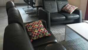 Un salon moderne avec un canapé en cuir noir, une table à manger avec des chaises, et un coin cuisine à l’arrière-plan.