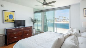 Ein Schlafzimmer mit einem Bett, einem Fernseher auf einer Kommode, einem Deckenventilator und einem großen Fenster mit Blick auf den Strand.