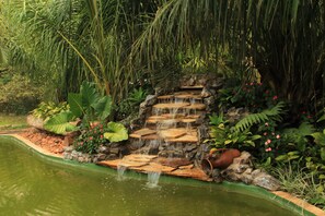 A stone waterfall in a lush garden with dense foliage and a pond. - Hotel Cabanas (Bonito)